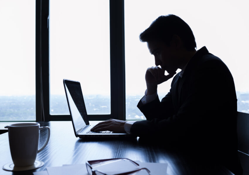 Silhouette of a man working at a laptop in an office with a window view, next to a coffee mug.